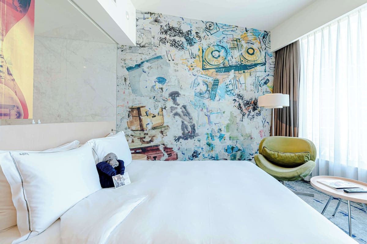 A hotel room with a large bed, a green armchair, a round coffee table, and a wall with abstract art.