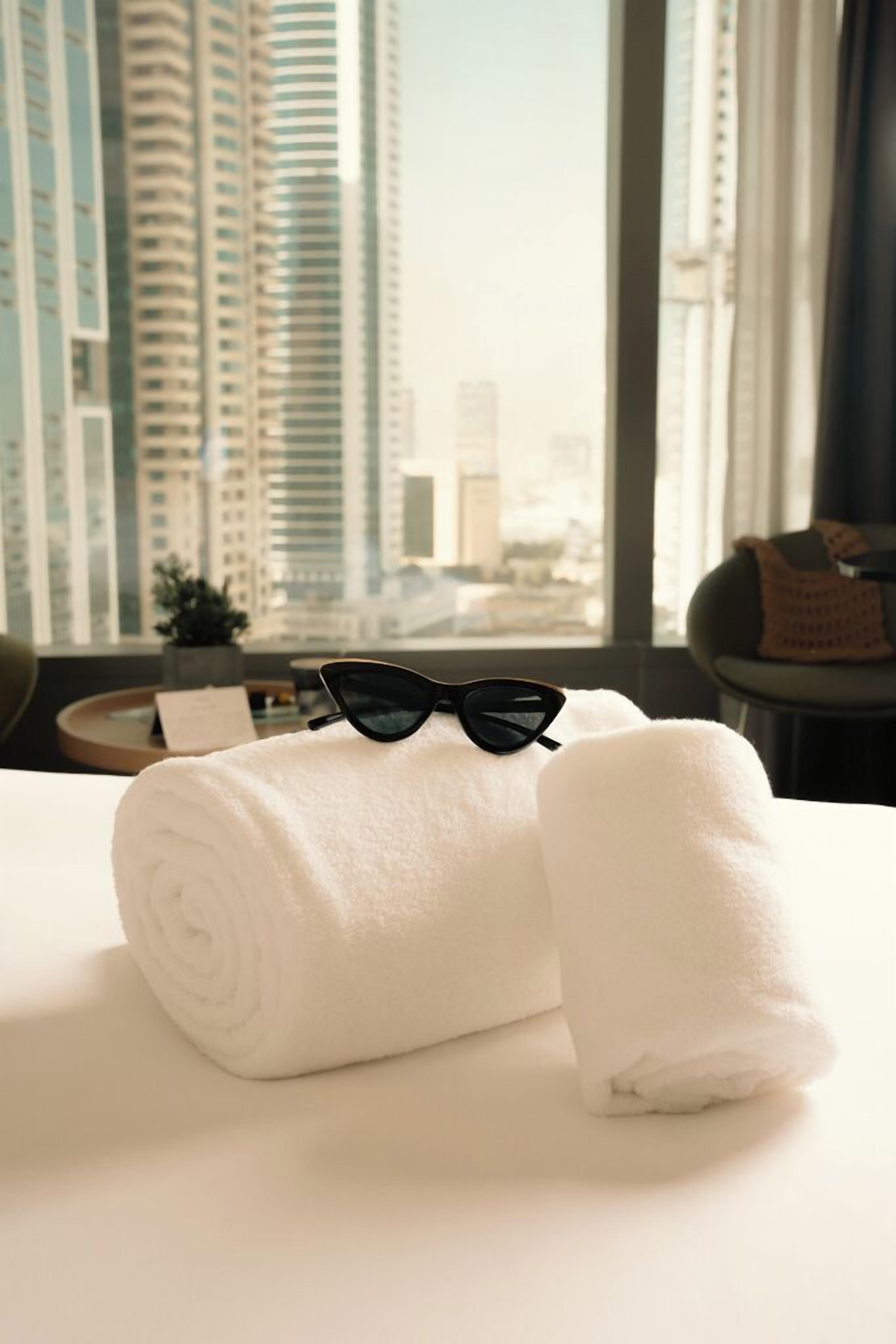 A neatly made bed with rolled white towels and black sunglasses on top, a small table with a plant, and a view of tall buildings through large windows.