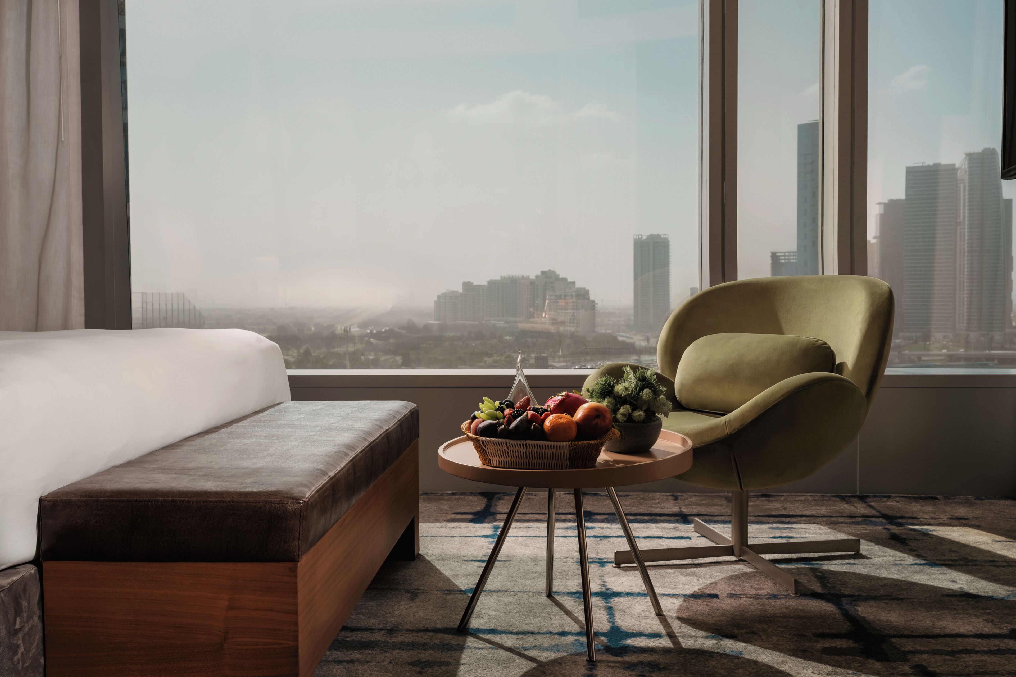 A modern hotel room with a bed, a green armchair, a round table with fruit, and a city view.
