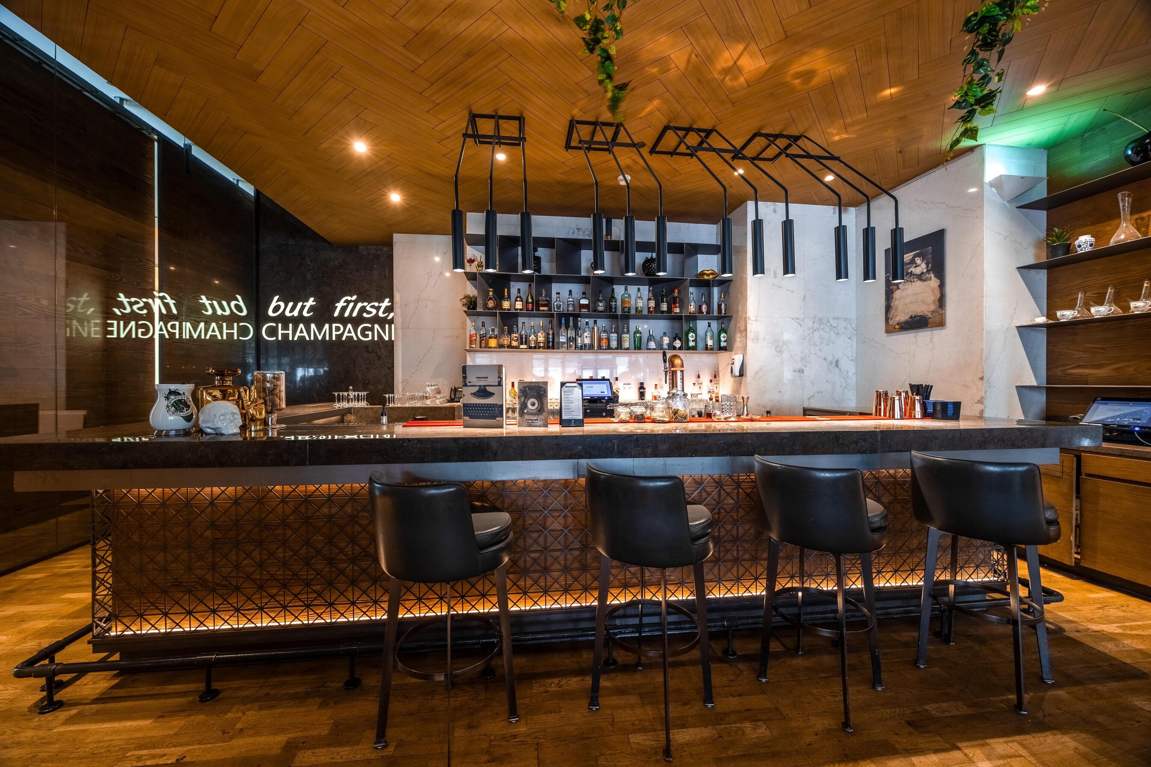A bar area with a wooden ceiling, bar stools, a counter with a cash register, and shelves with bottles.