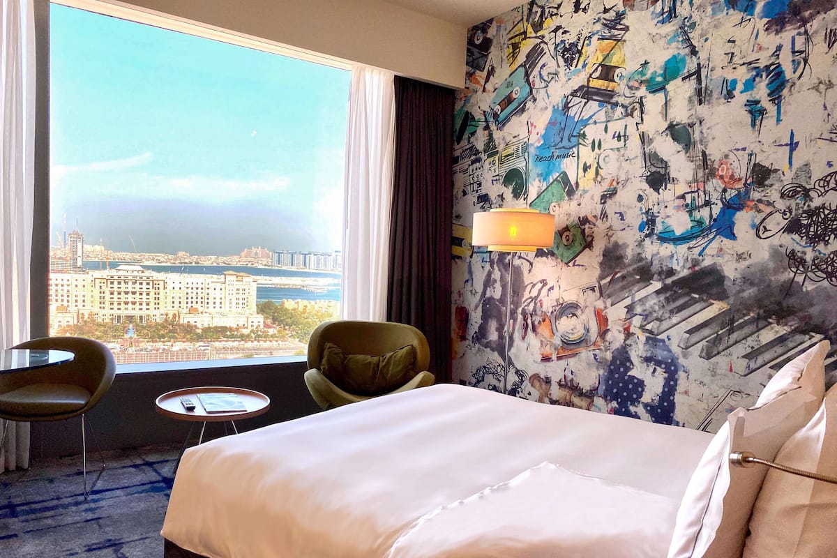 A hotel room with a large window, a bed, a chair, a small table, and a wall with a colorful abstract design.