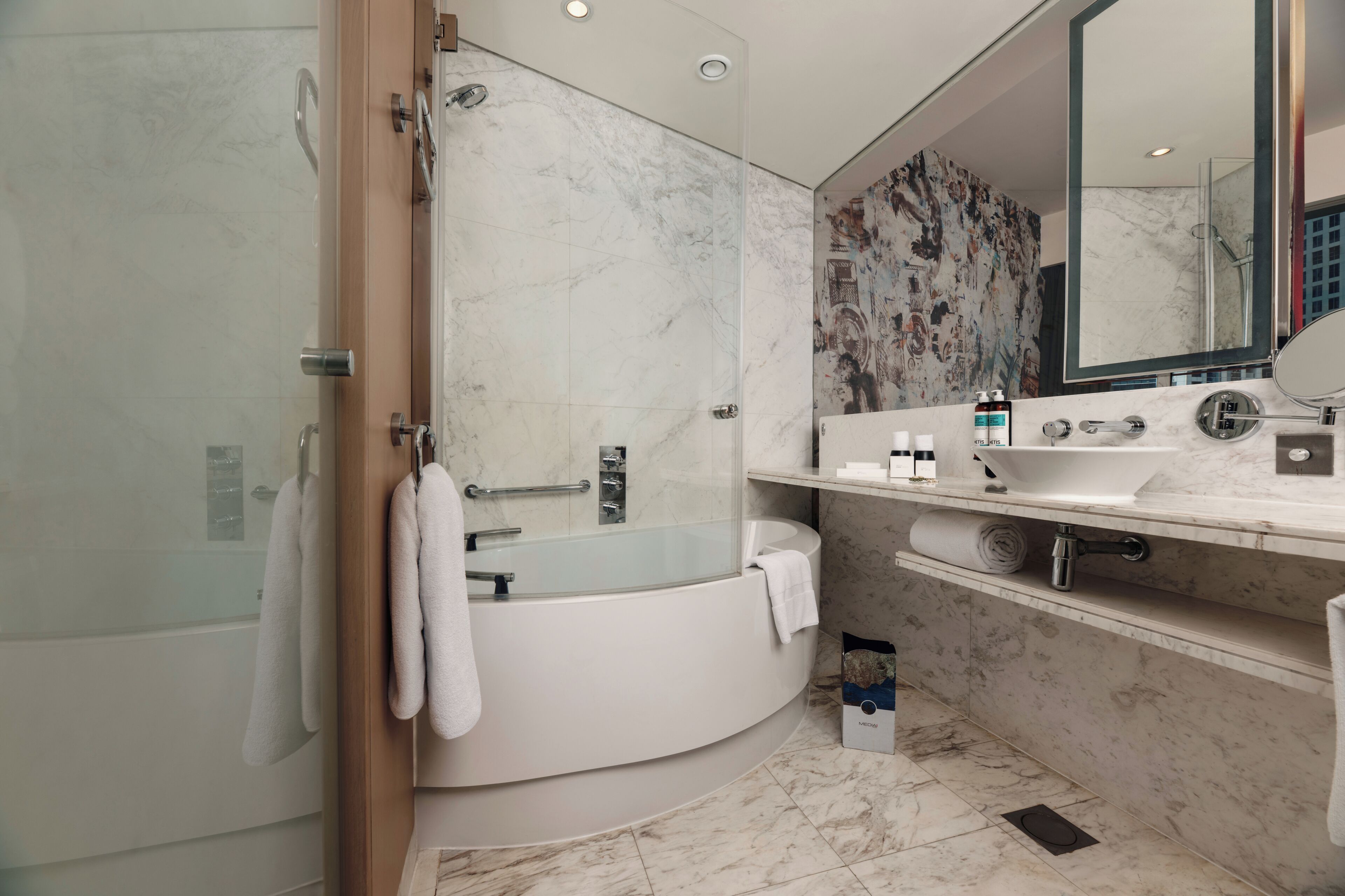 A modern bathroom with a glass-enclosed shower, a freestanding bathtub, a large mirror, and a marble-tiled wall.