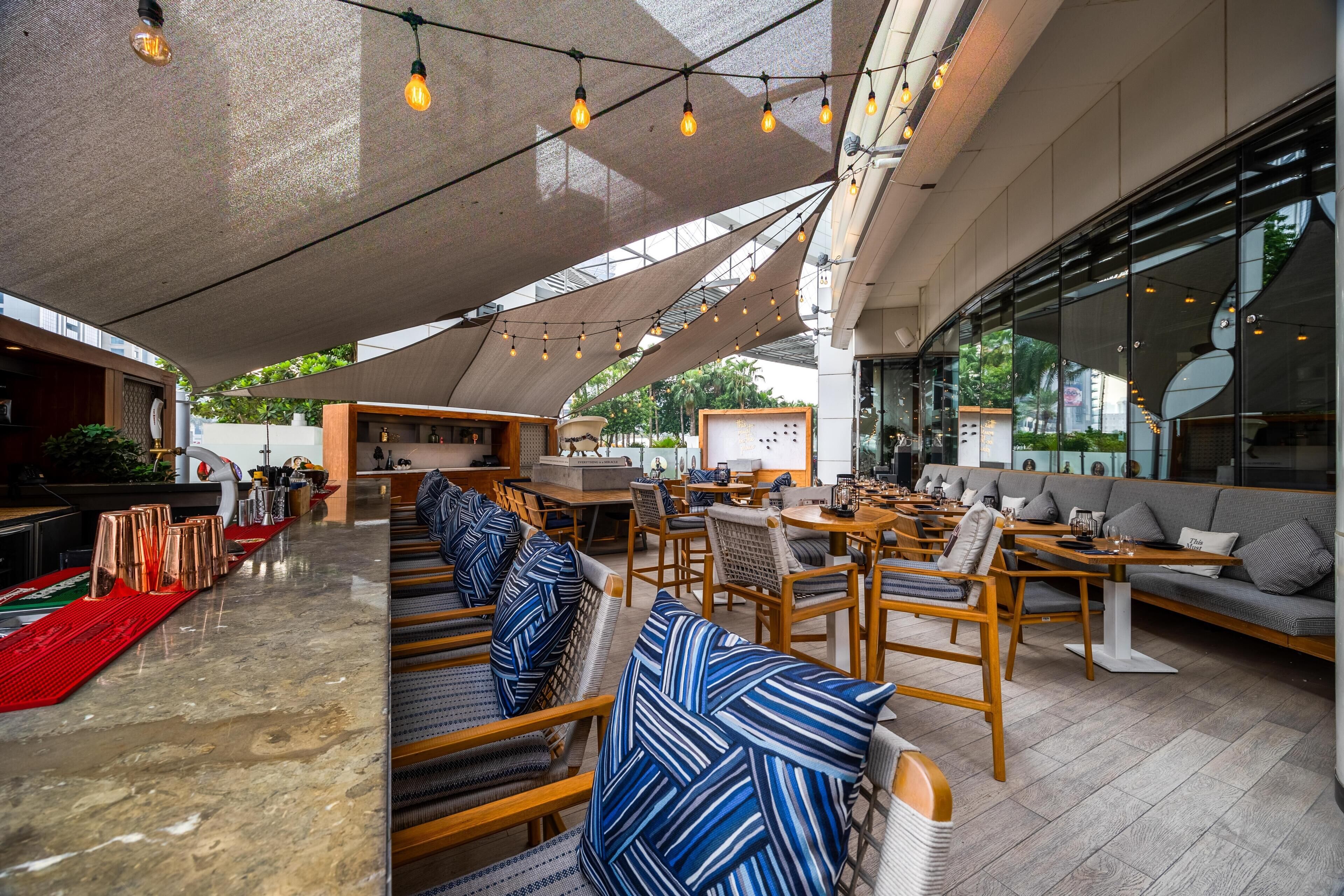 A modern outdoor seating area with wooden tables and chairs, a bar with copper mugs, and a large window offering views of the surrounding area.