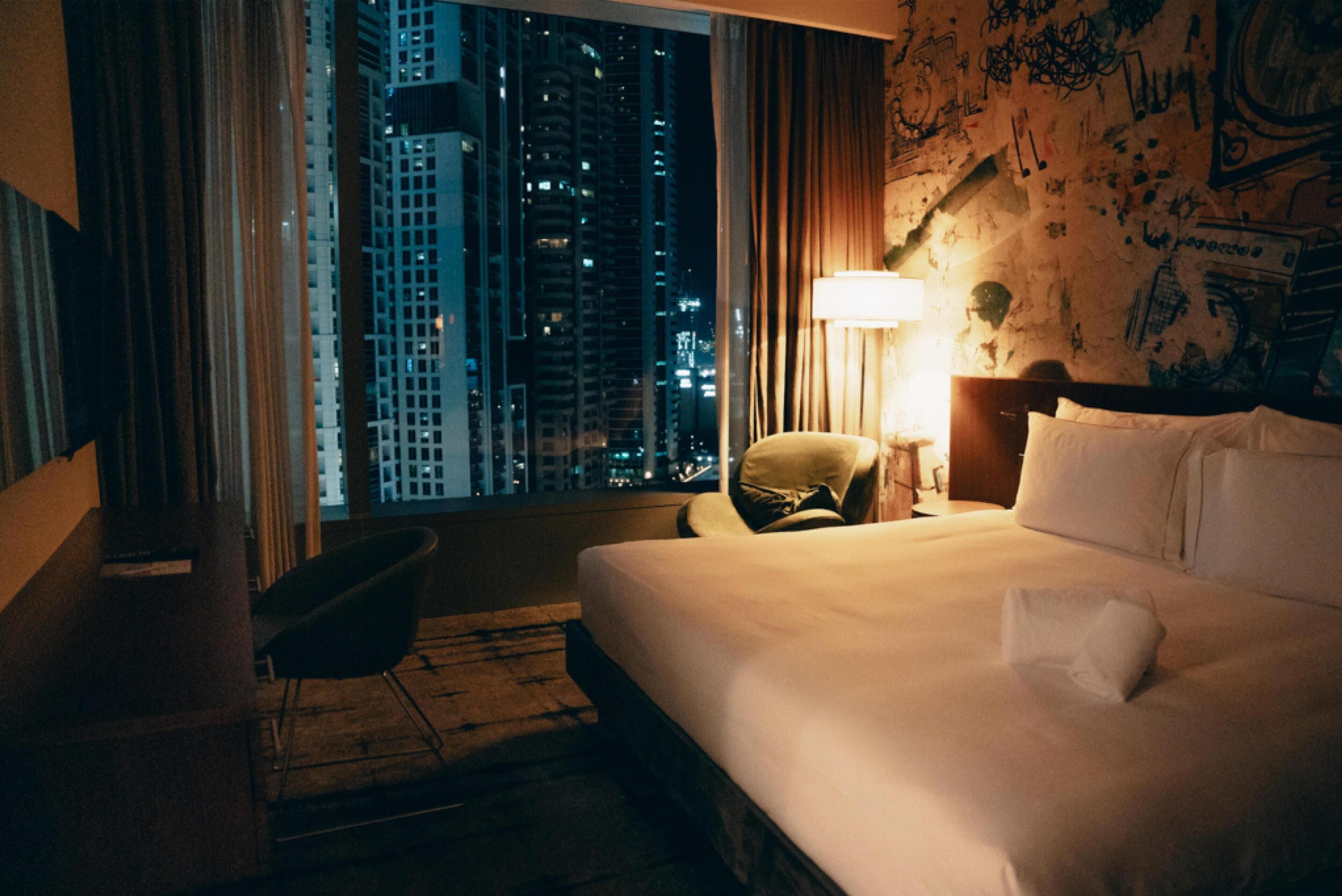 A hotel room with a bed, a chair, a nightstand, a lamp, and a view of city buildings.