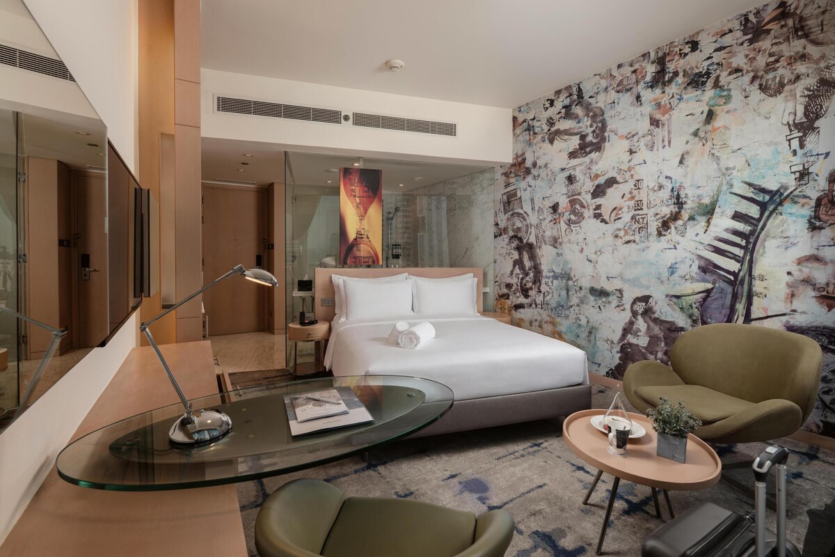 A modern hotel room with a glass bed, a glass coffee table, a green armchair, a small side table with a plant, and a large abstract mural on the wall.