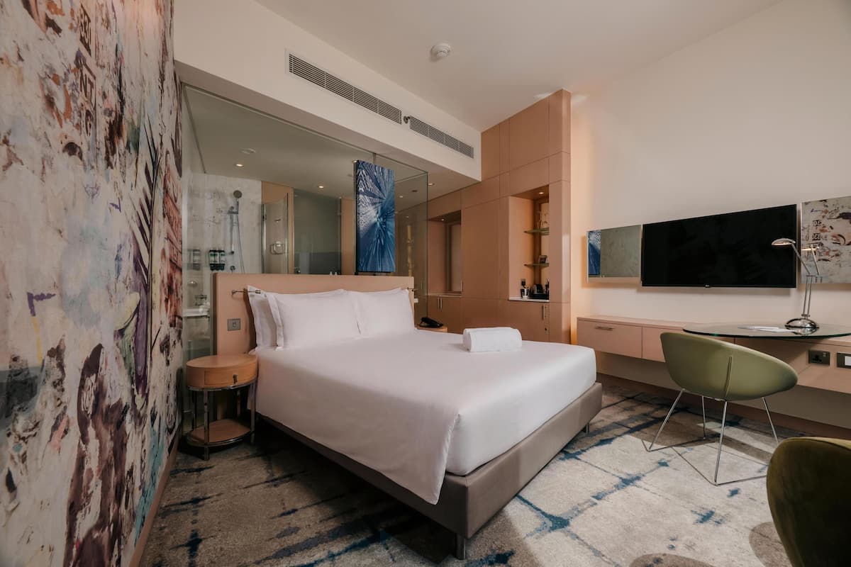 A modern hotel room with a large bed, a desk, a chair, a TV, and a bathroom visible through a glass partition.
