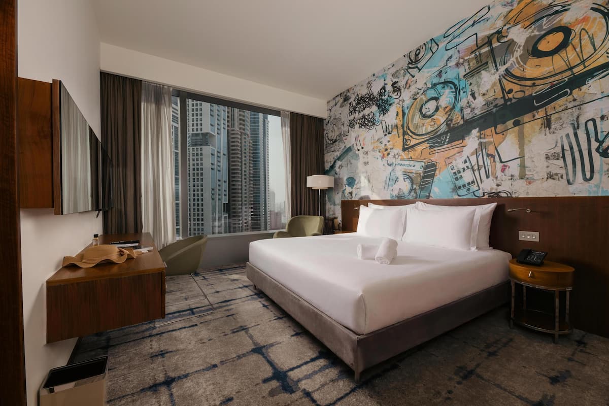 A hotel room with a large bed, a desk, a chair, a lamp, and a view of tall buildings.