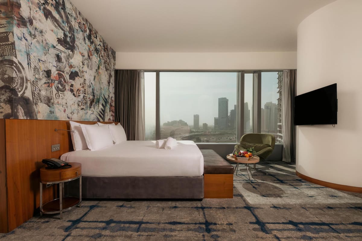 A hotel room with a large bed, a city view, a flat-screen TV, a green armchair, and a small table with fruit.