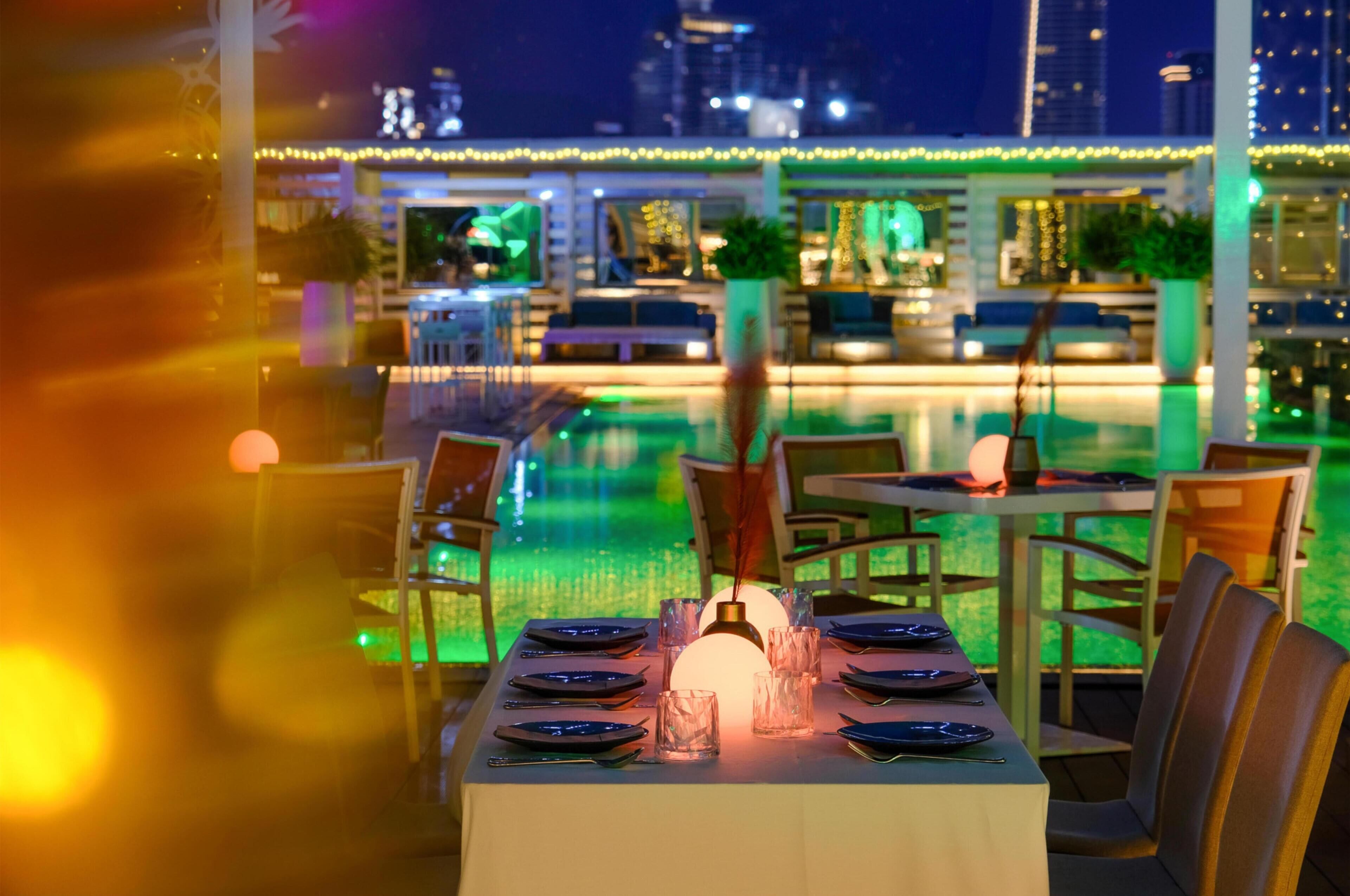 Comedor con mesas dispuestas para dos, una piscina al fondo y vistas a la ciudad.