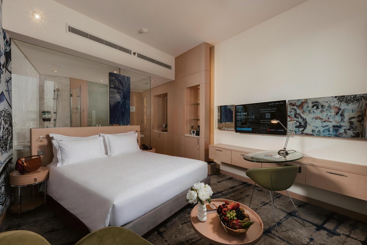A modern hotel room with a large bed, a desk with a computer, a TV, and a glass shower enclosure.