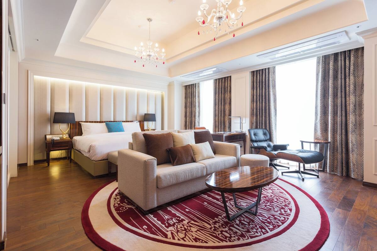 A spacious hotel room with a large bed, a sofa, a coffee table, and a desk. There is a chandelier, curtains, and a view of the outside.