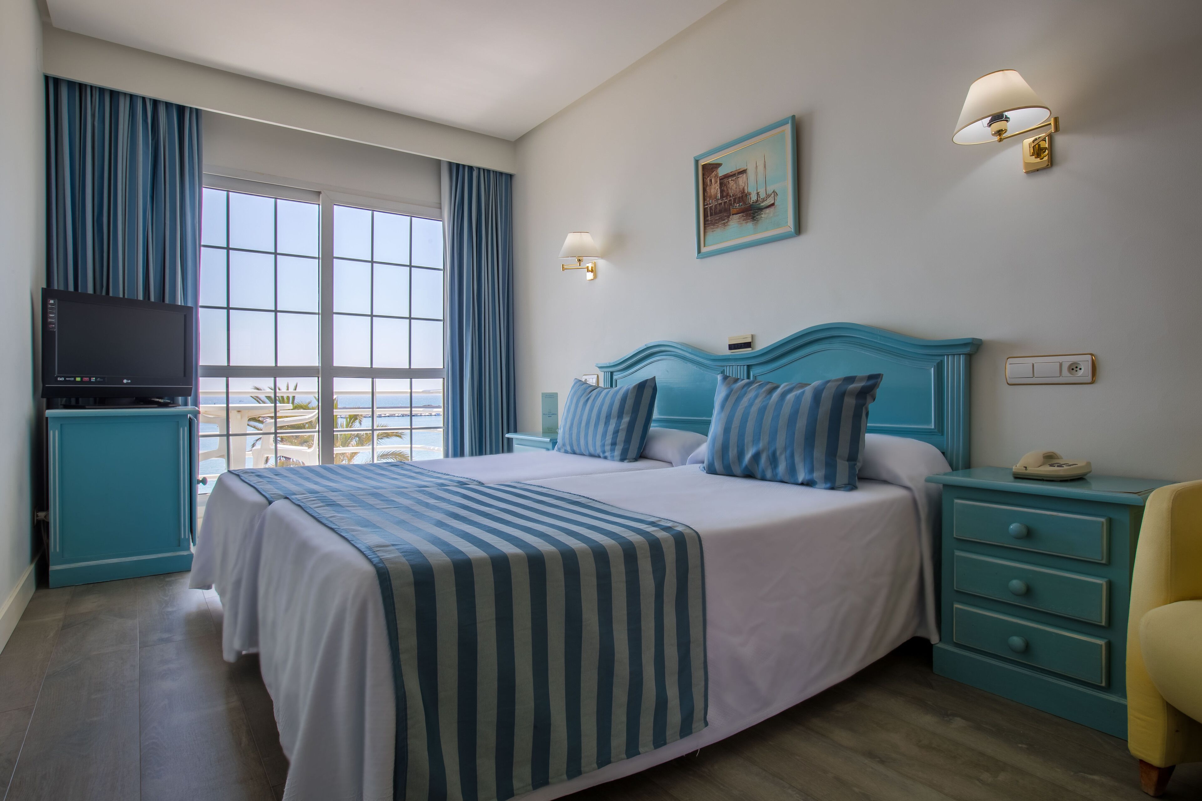 A hotel room with a large bed, a television, a nightstand, and a view of the sea.