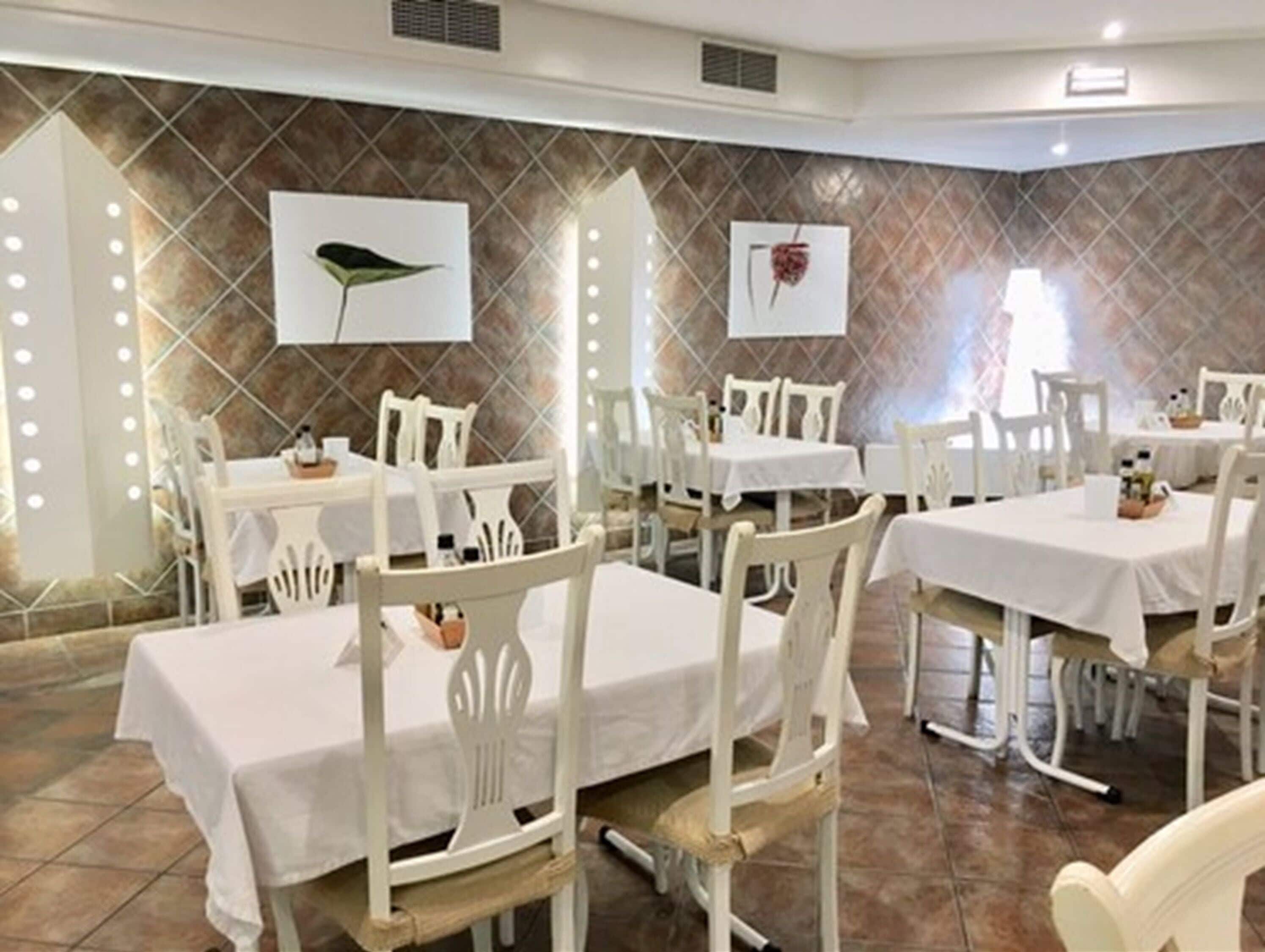 Un restaurant avec des tables et des chaises blanches, un mur recouvert de carreaux orné d’œuvres d’art, et une atmosphère chaleureuse.