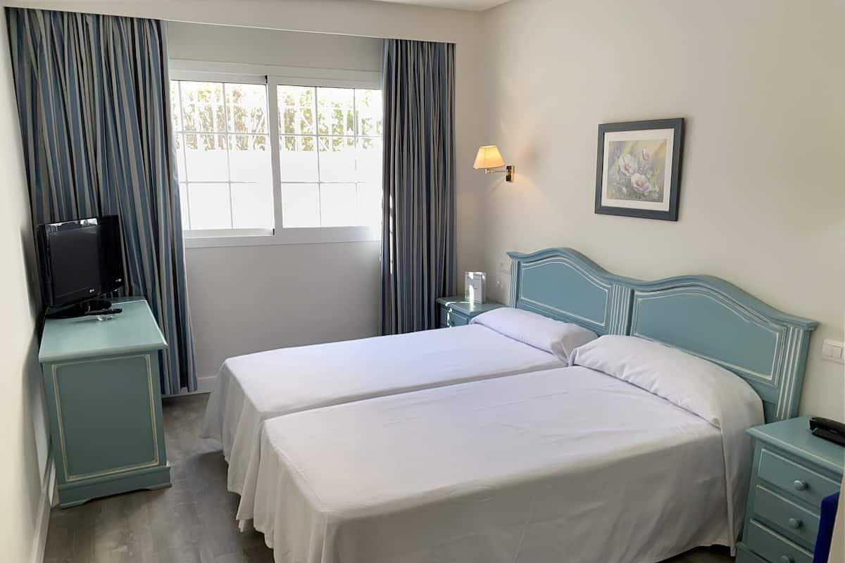 Una habitación de hotel con dos camas, un televisor sobre una mesita auxiliar, una lámpara de noche y un cuadro en la pared.