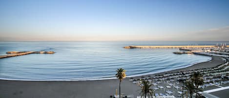 A beach with a curved walkway, palm trees, and a marina with boats.