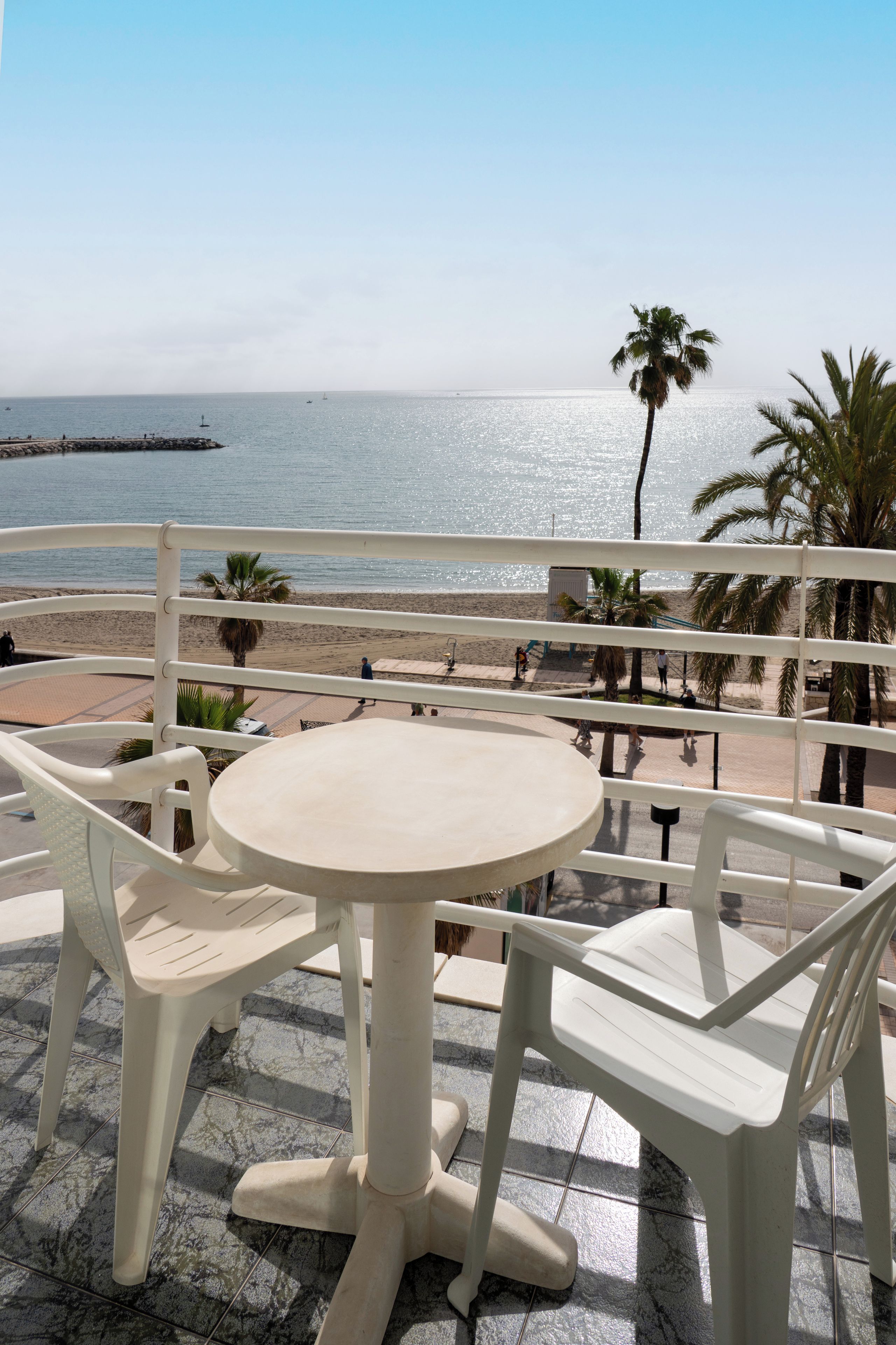 A terrace with a round table and white chairs, overlooking a beach with palm trees and a clear sky.