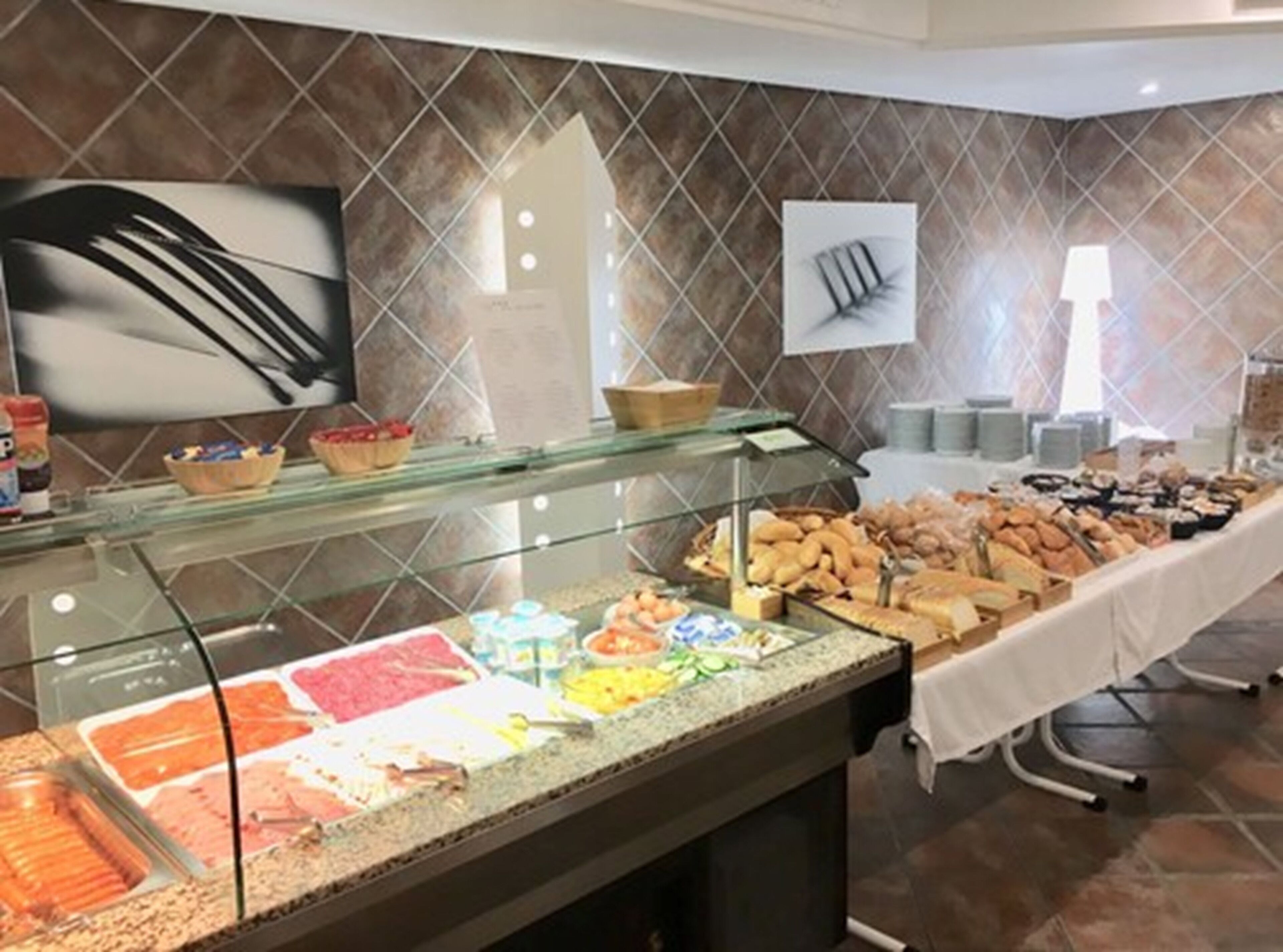 A buffet area with various food options, including meats, cheeses, and pastries, in a modern setting with tiled walls and a glass display case.