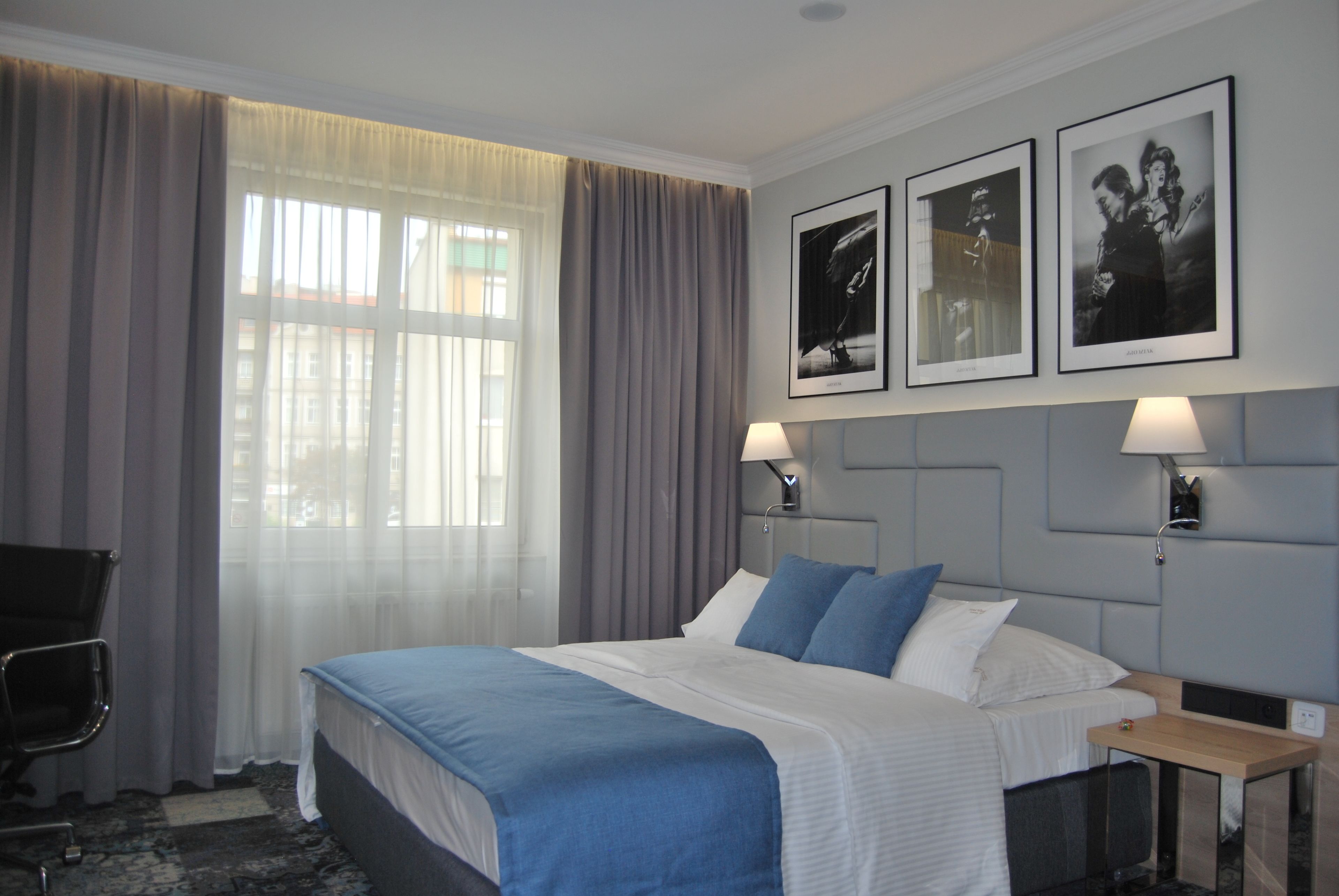A hotel room with a bed, two bedside lamps, a desk, and a chair. There are curtains on the window and two framed pictures on the wall.