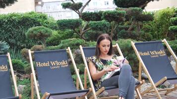 A woman sitting on a lounge chair with a book, surrounded by well-maintained bushes and trees, in an outdoor setting.