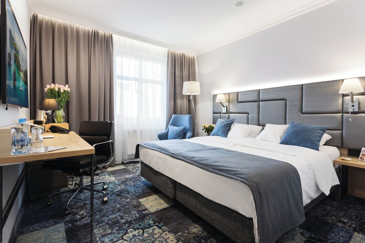 Habitación de hotel con una cama grande, un escritorio con silla, una lámpara y un ventanal con cortinas.