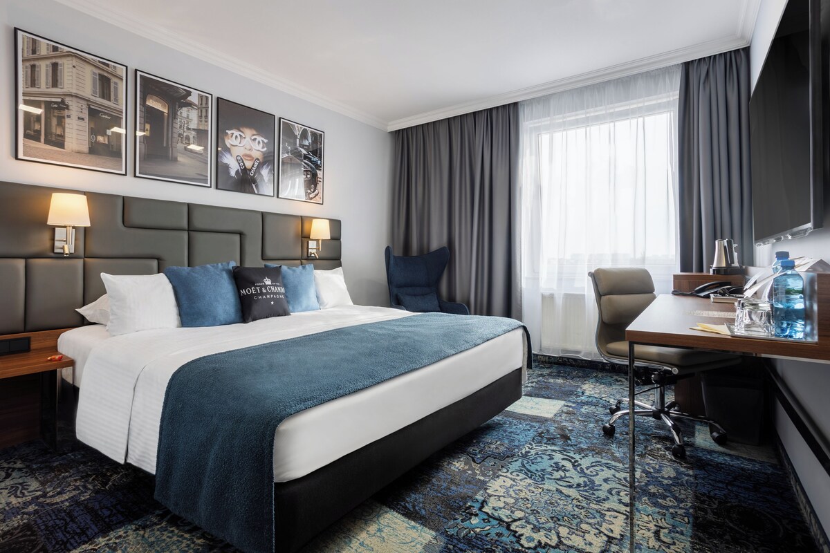 Habitación de hotel con una cama grande, un escritorio, una silla, un televisor y obras de arte en la pared.