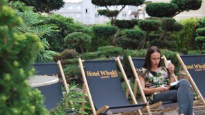 Una mujer sentada en una silla de descanso con un cartel que dice 'Hotel Wloski', rodeada de arbustos y árboles bien cuidados.