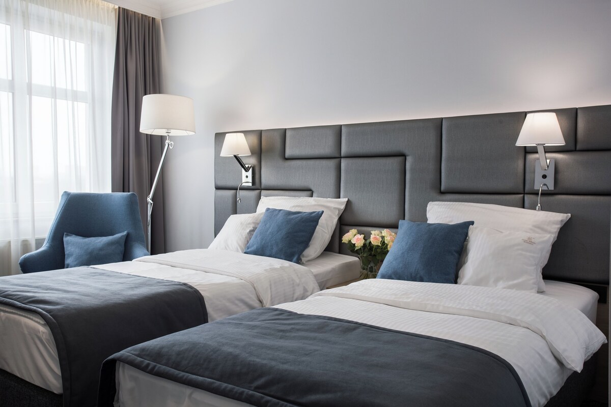 Habitación de hotel con dos camas, un ventanal con cortinas y una lámpara moderna.