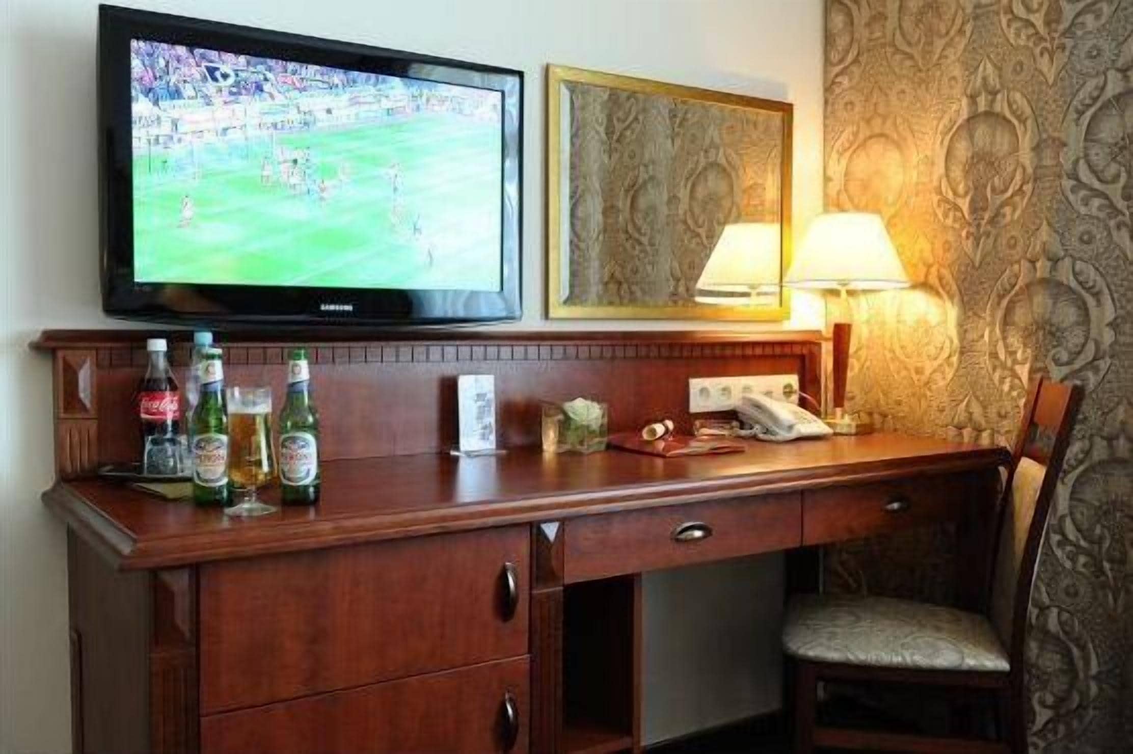 A hotel room with a wooden desk, a television, a lamp, and a patterned wallpaper.