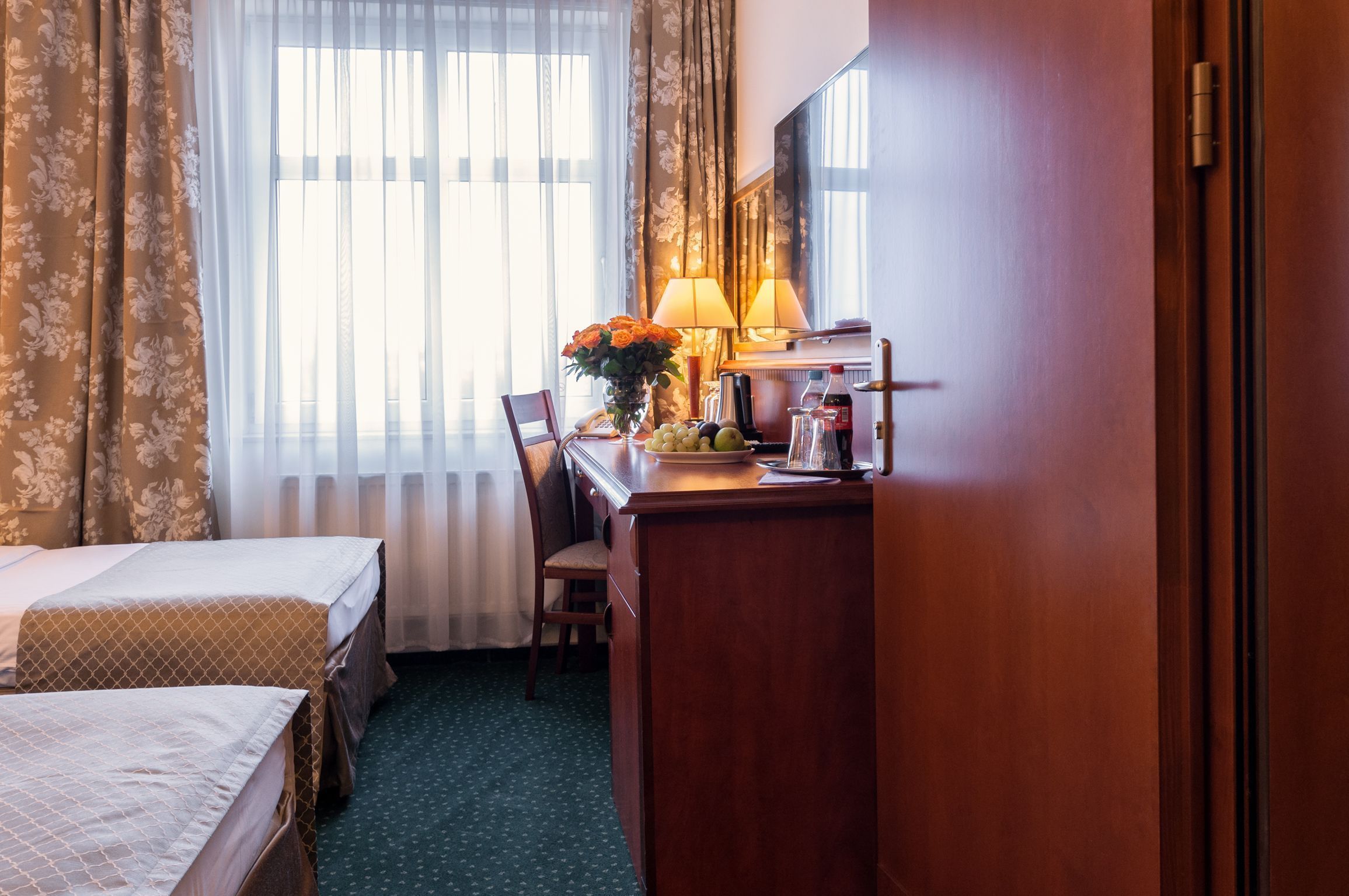 A hotel room with two beds, a desk with a lamp, a chair, and a view of the outdoors through sheer curtains.