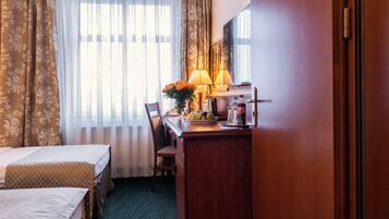 A hotel room with two beds, a desk with a lamp, a chair, and a view of the outdoors through sheer curtains.