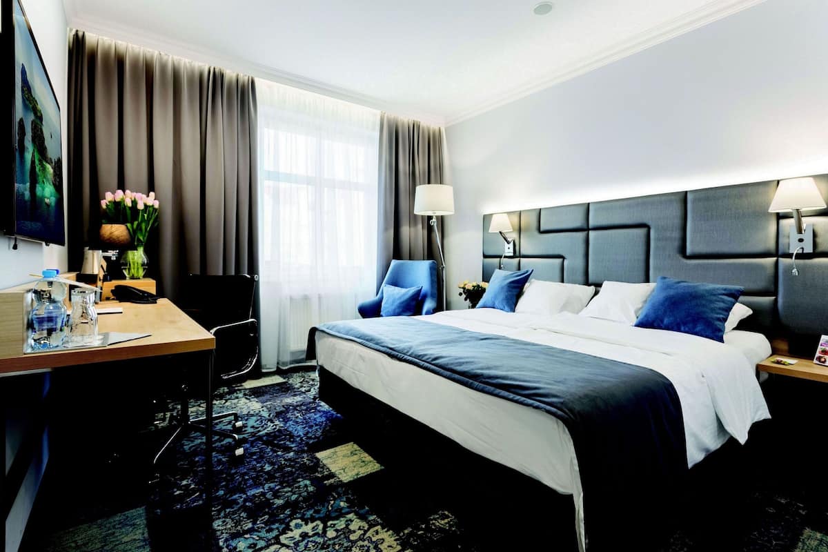 A hotel room with a large bed, a desk with a chair, a TV, and a window with curtains.