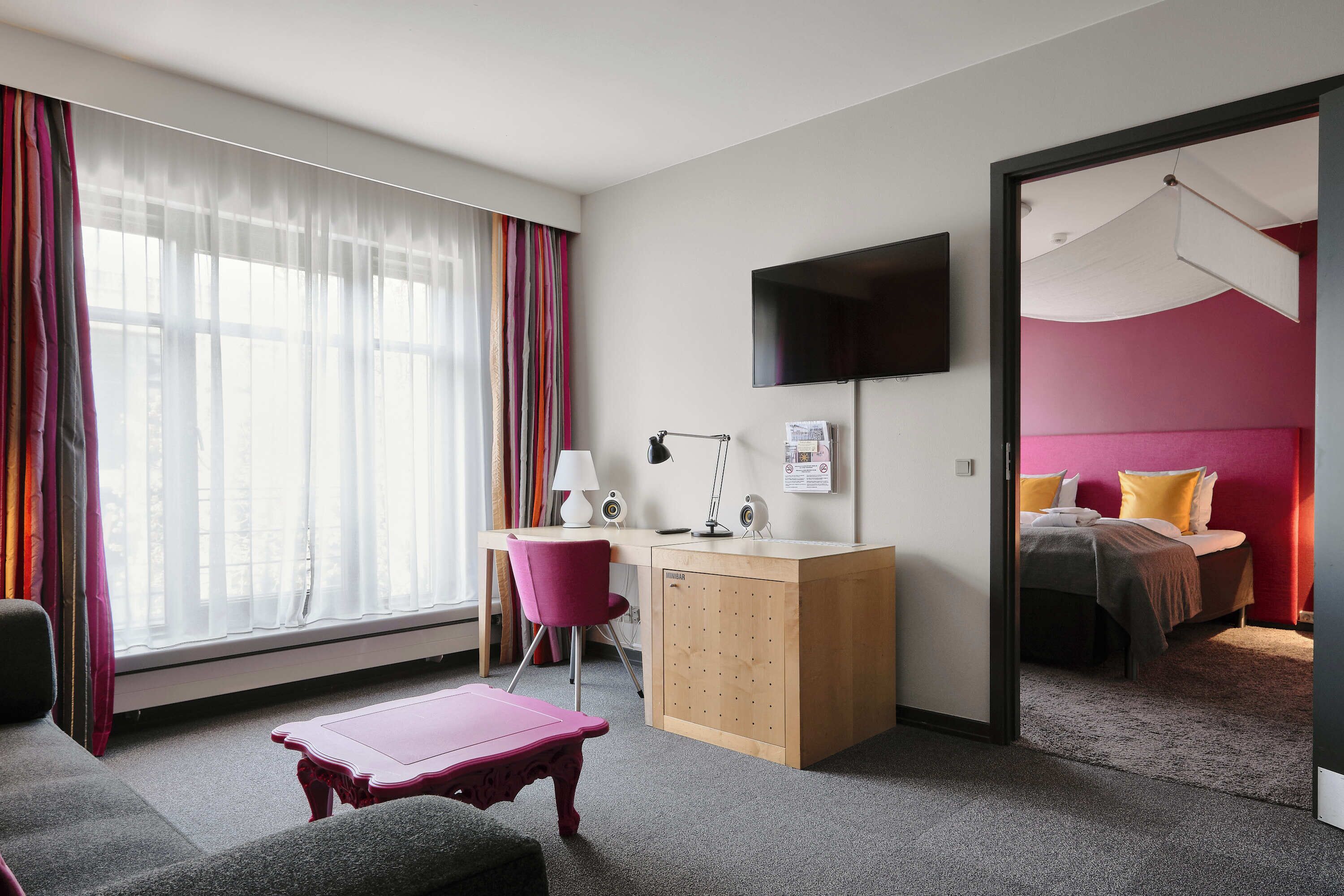 A hotel room with a desk, a chair, a bed, a TV, and a window with curtains.