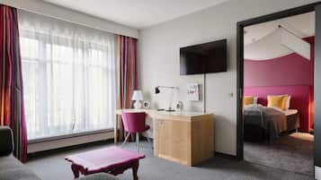 Ein Hotelzimmer mit Schreibtisch, Stuhl, Bett, Fernseher und einem Fenster mit Vorhängen.