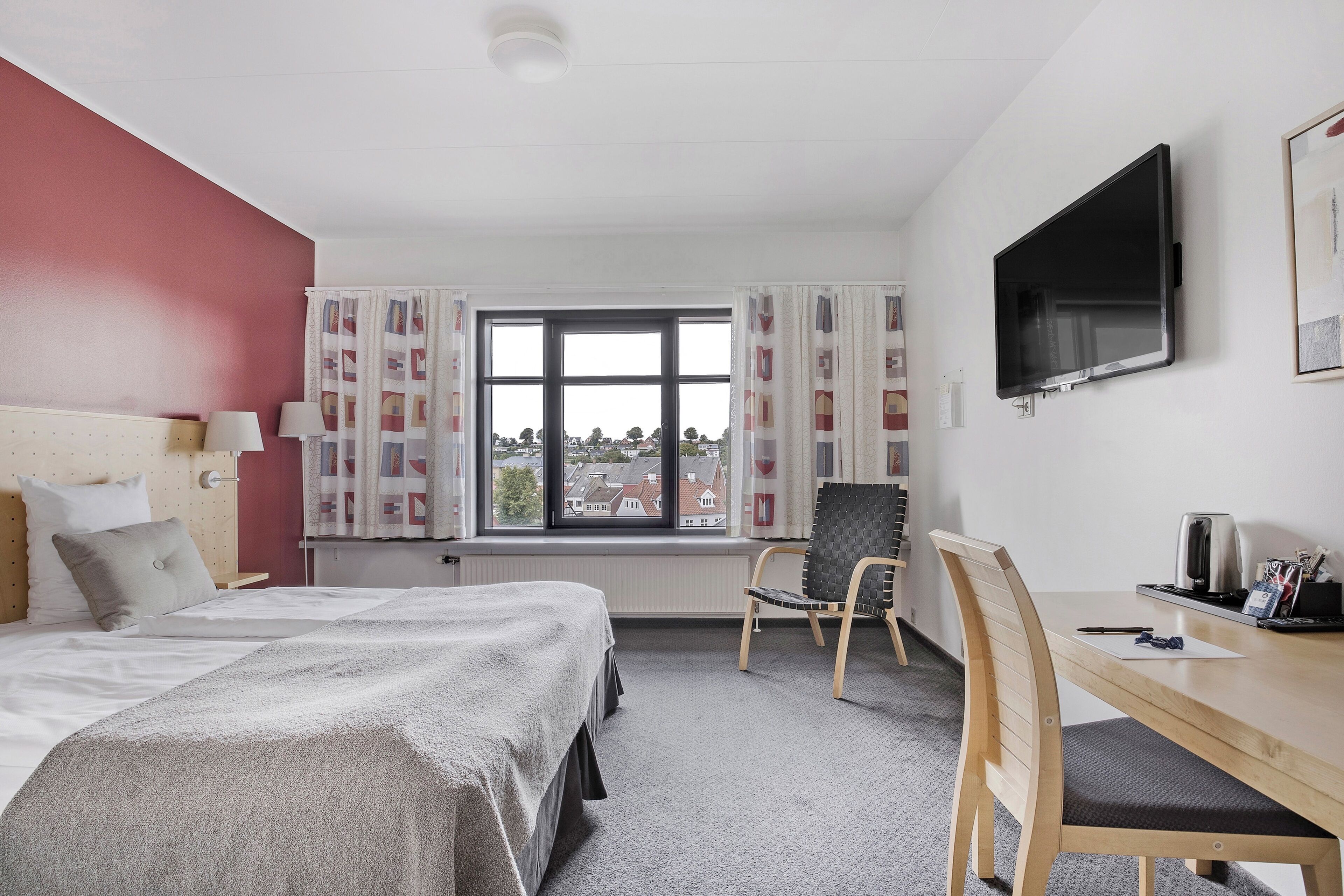 Et hotelværelse med seng, skrivebord, stol, fjernsyn og et vindue med gardiner.