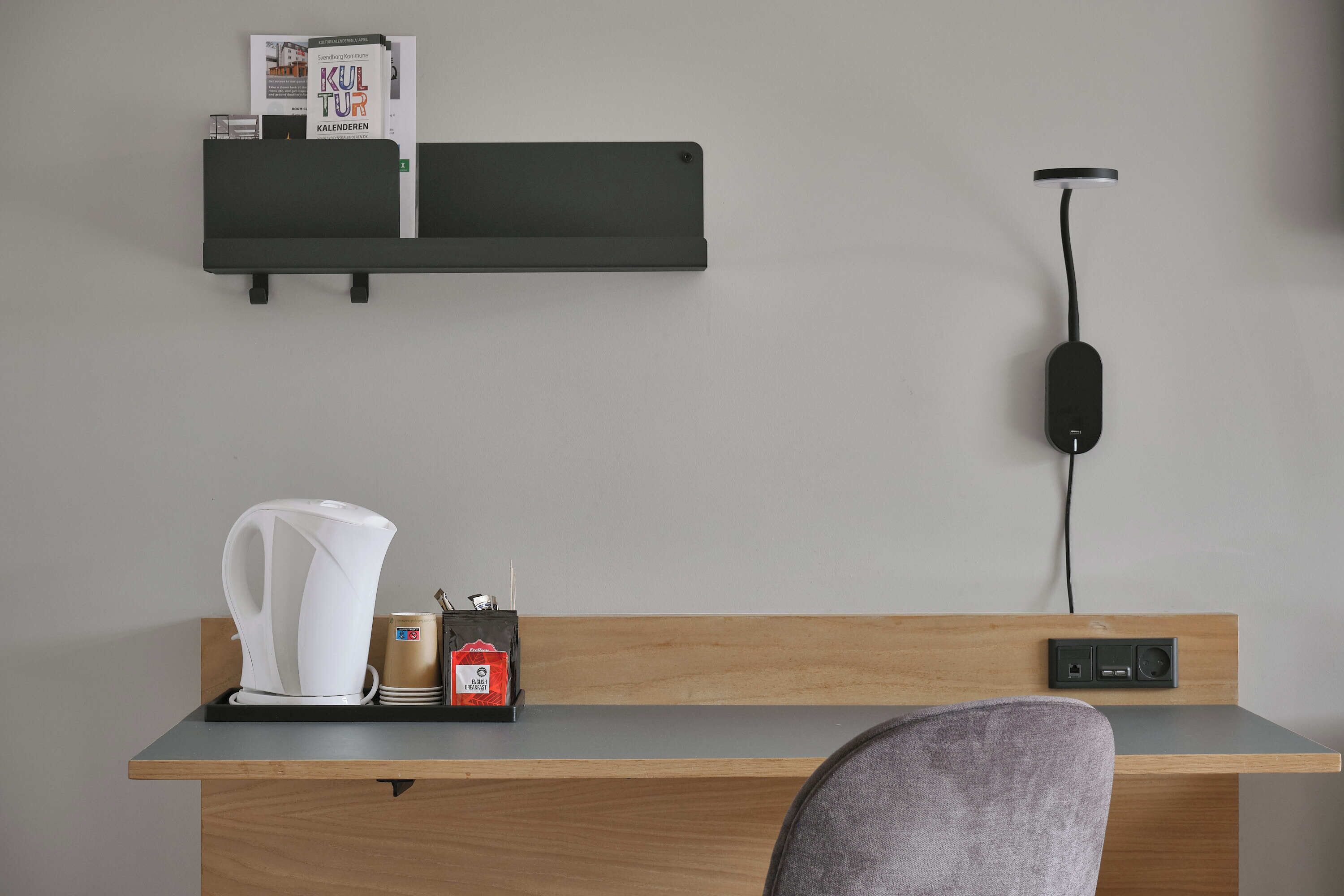 A modern hotel room with a wooden desk, a black charging station, a white kettle, and a coffee maker.