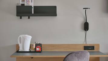 A modern hotel room with a wooden desk, a black charging station, a white kettle, and a coffee maker.