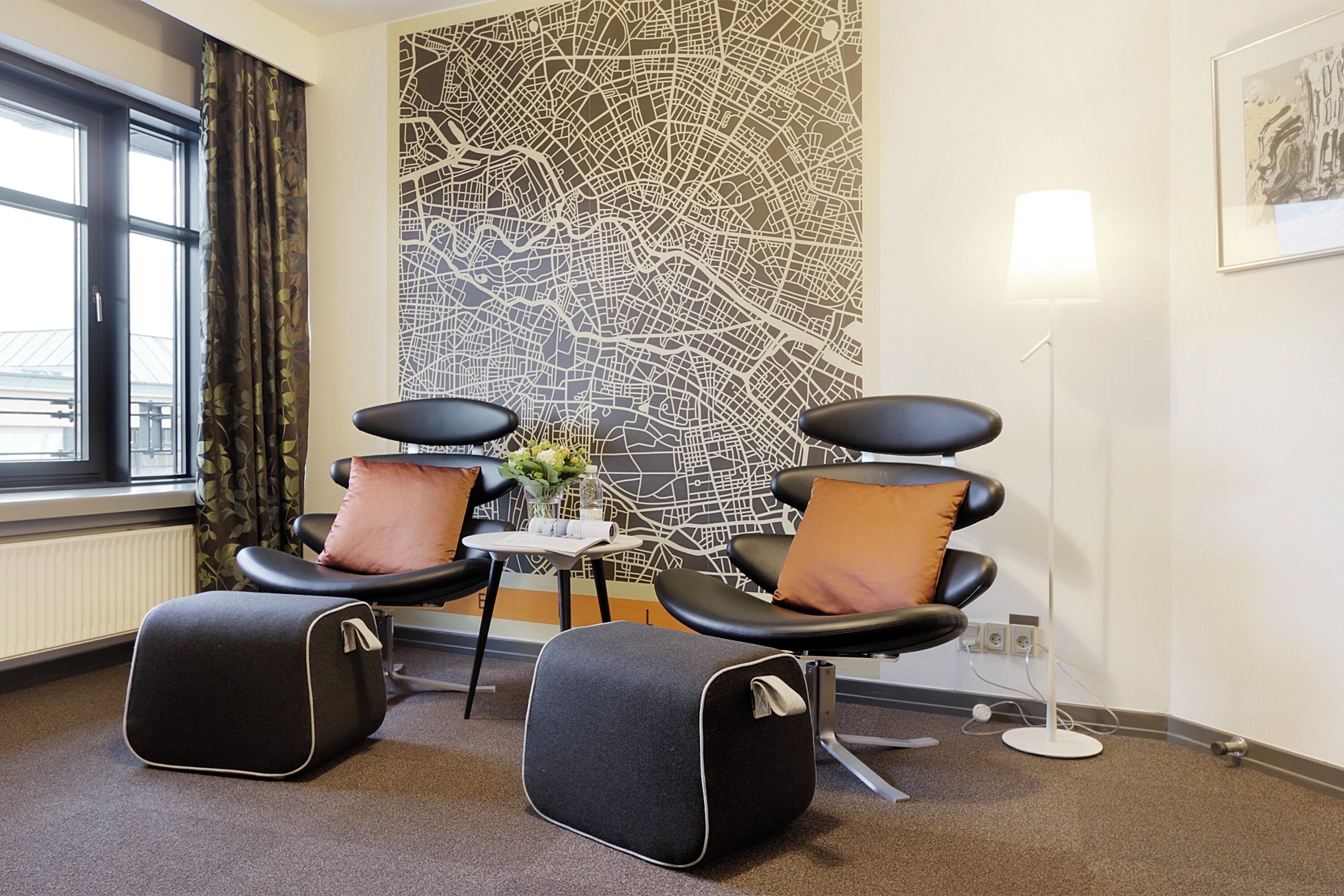 A modern waiting area with two black chairs, a small table with a vase, and a large abstract wall art piece.