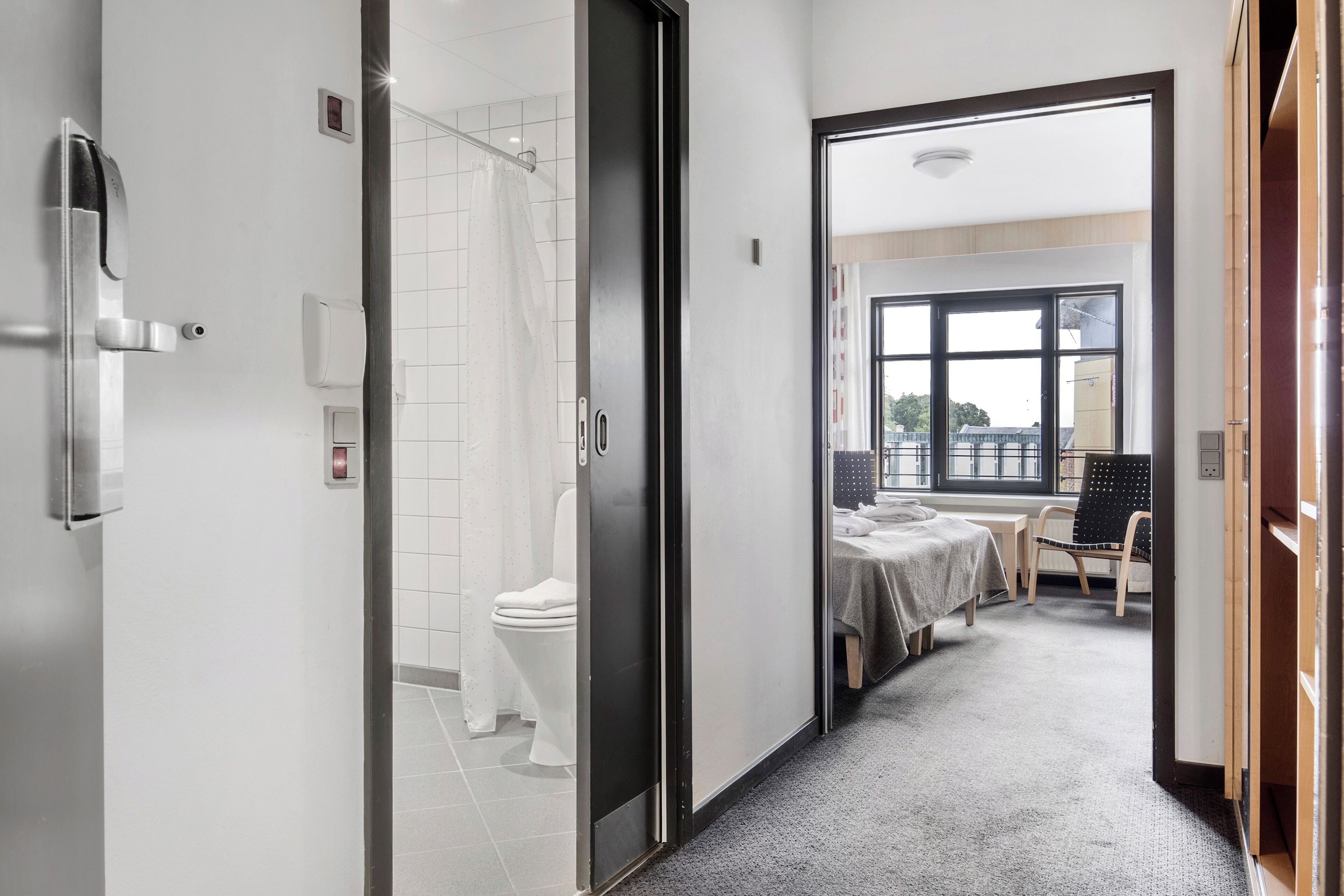 Une chambre d’hôtel équipée d’une douche, de toilettes, d’un lit et offrant une vue sur la ville grâce à de grandes fenêtres.