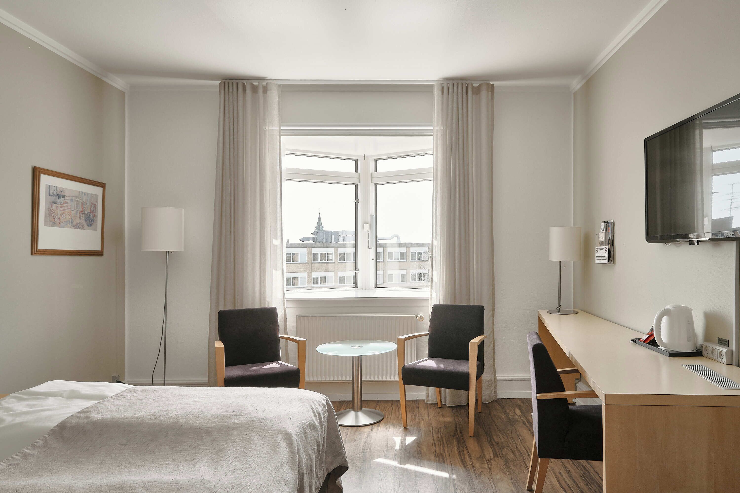 A hotel room with a bed, two armchairs, a small round table, a desk with a computer, a TV, and a window with curtains.