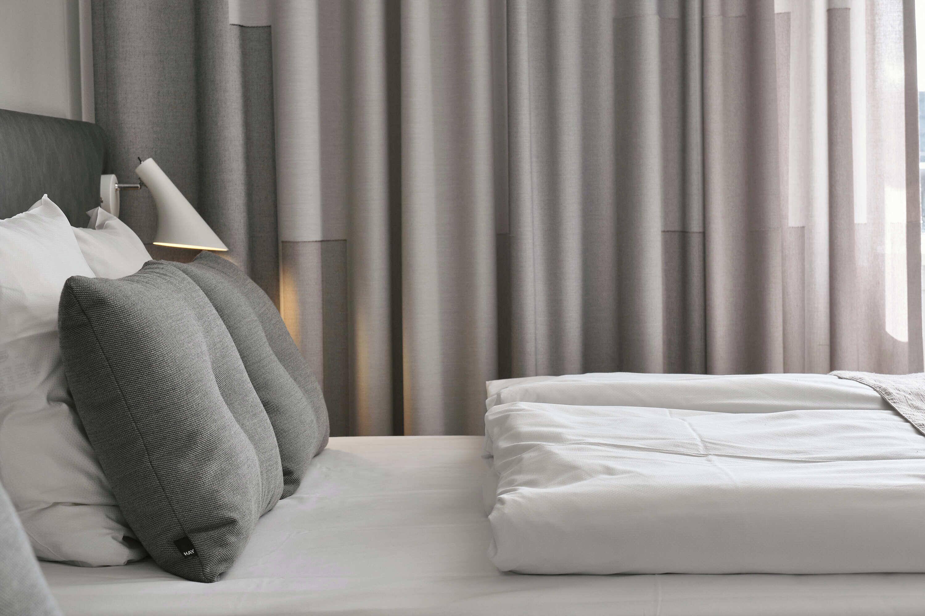 A neatly made bed with white bedding and gray pillows, a bedside lamp, and a view of curtains and a window.