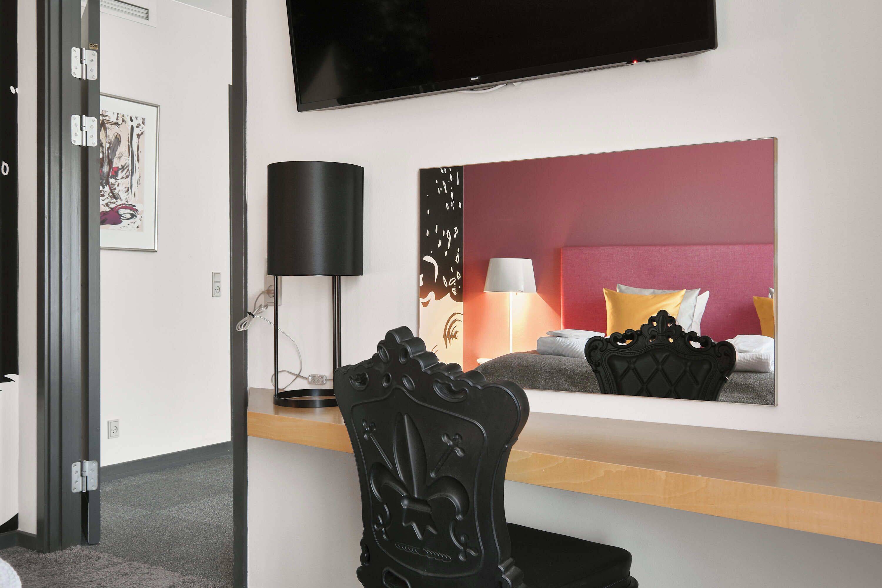 Une chambre d’hôtel moderne dotée d’un téléviseur à écran plat fixé au mur, d’une table de chevet avec une lampe, d’une tête de lit décorative et d’une œuvre d’art encadrée accrochée au mur.