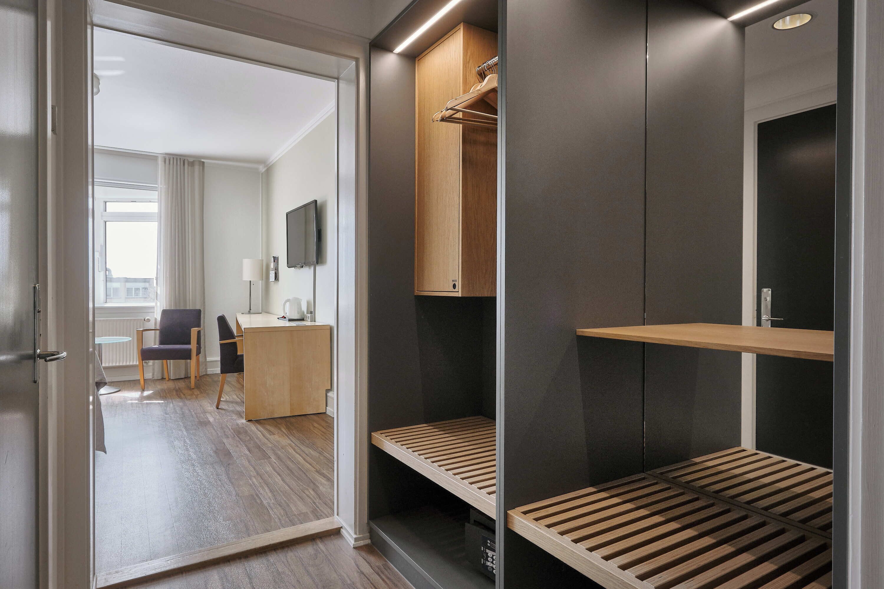 Una habitación de hotel moderna con un escritorio de madera, una silla, un televisor y un vestidor con estantes y espacio para colgar.