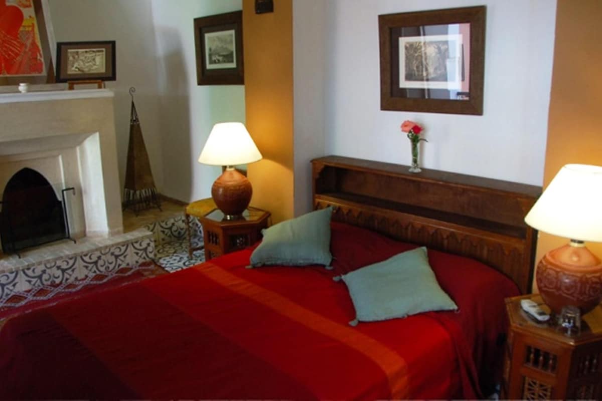 A hotel room with a fireplace, two bedside lamps, a bed with red bedding, and a wooden headboard.