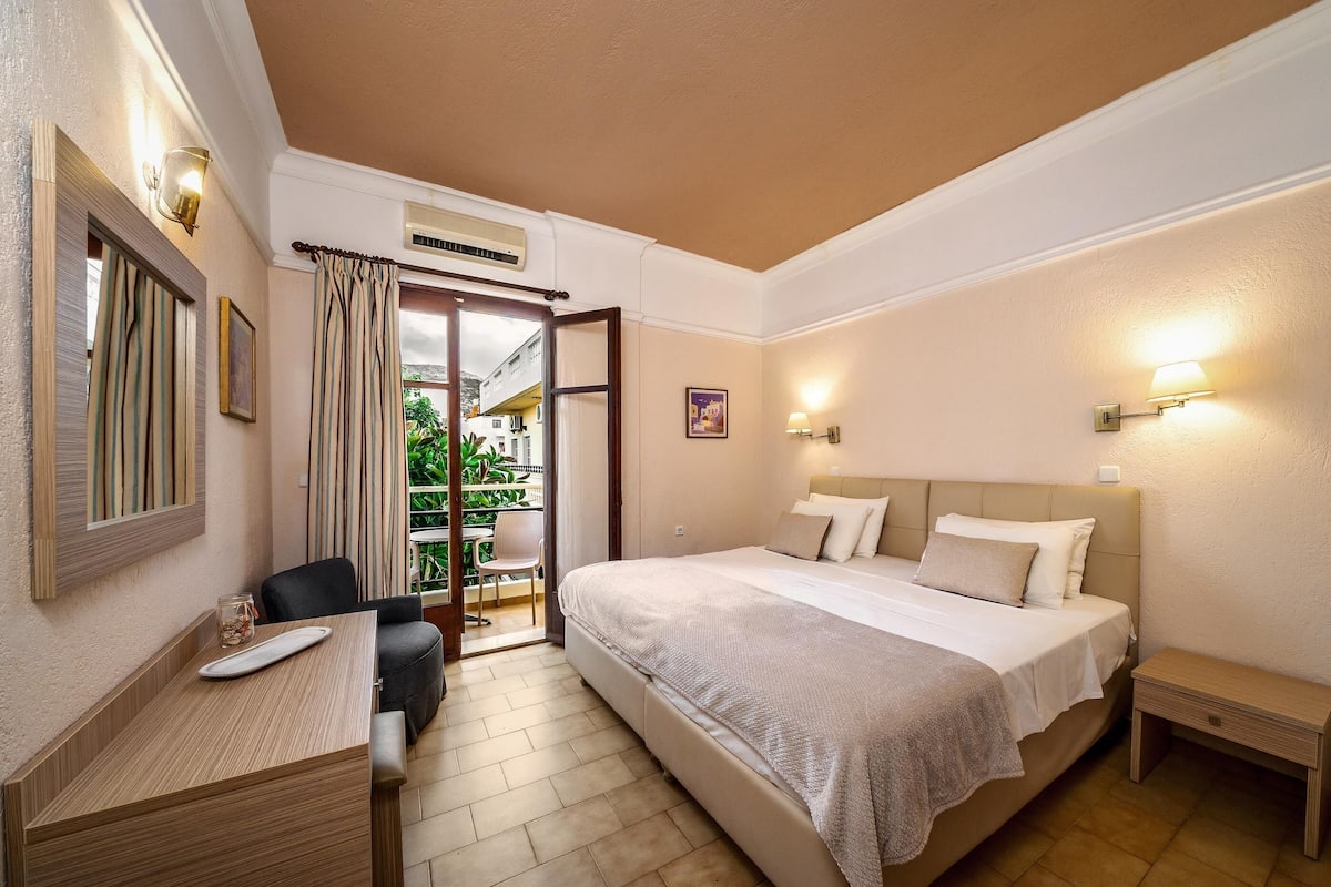A hotel room with a bed, bedside table, desk with chair, and a view of the outside balcony.