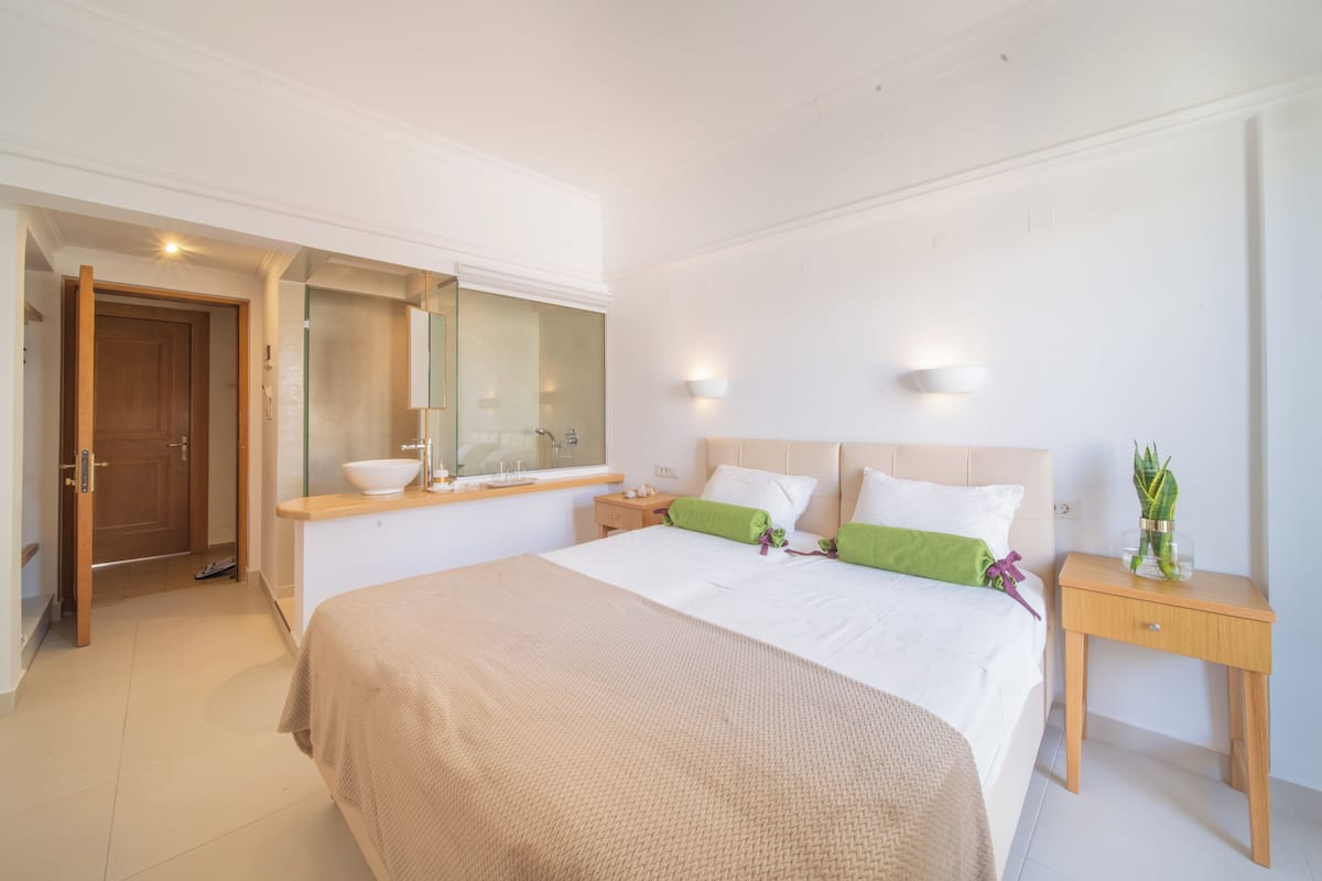 A modern bedroom with a large bed, a wooden nightstand, a mirror, and a small window.