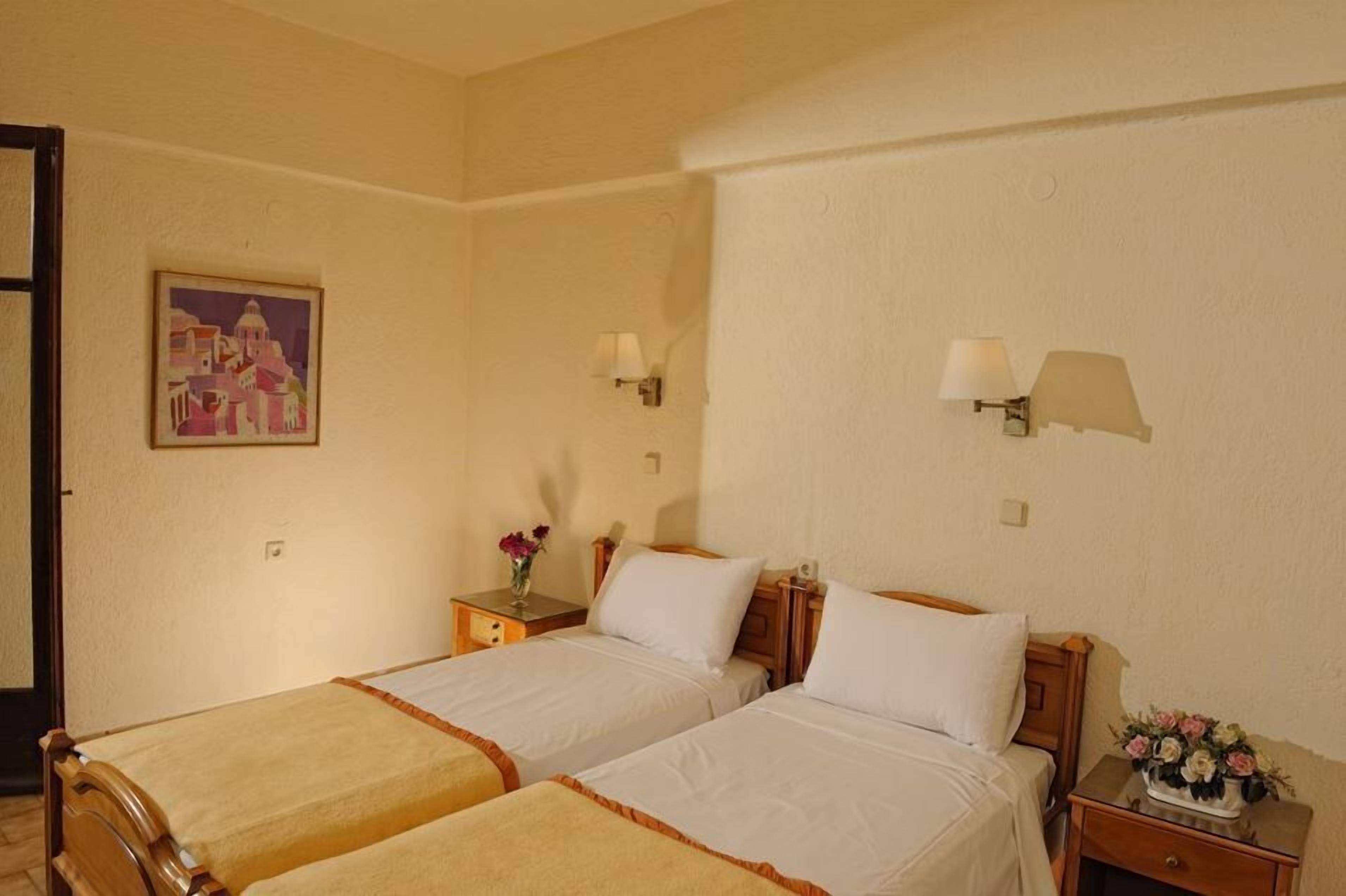 Een hotelkamer met twee eenpersoonsbedden, een nachtkastje met een lamp, een schilderij aan de muur en een vaas met bloemen op het nachtkastje.