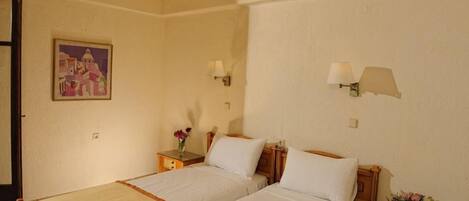 A hotel room with two single beds, a nightstand with a lamp, a painting on the wall, and a vase with flowers on the nightstand.