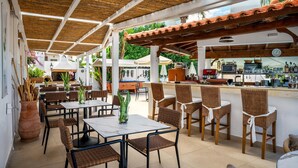 An outdoor café with tables and wicker chairs, a bar area with kitchen appliances, and a tiled floor. - Malia Mare (Hersonissos)