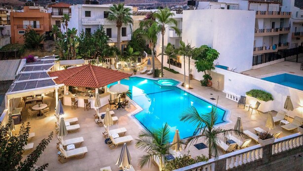 A rooftop pool area with lounge chairs and umbrellas, surrounded by buildings and palm trees. - Malia Mare (Hersonissos)