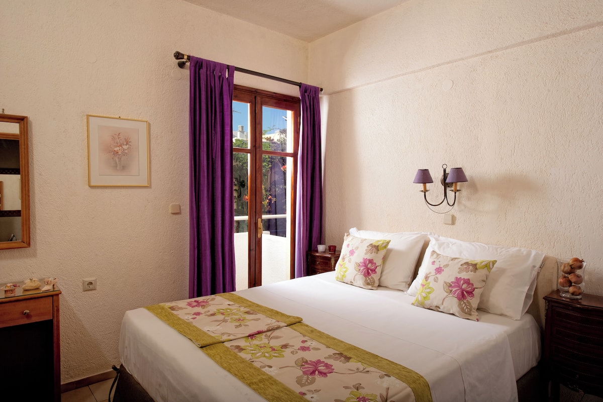 A hotel room with a bed, a nightstand, a mirror, a window with purple curtains, and a wall-mounted lamp.